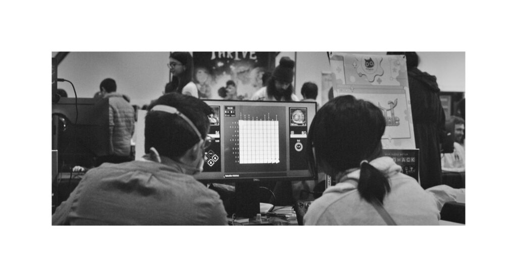 A monochrome photo of Philip showing someone how to play GRIDHACK on a monitor screen.